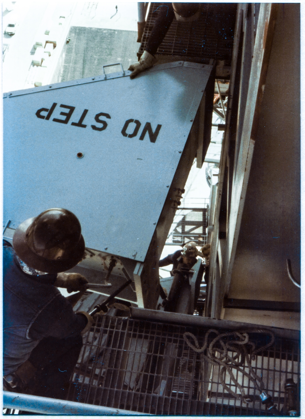 Image 113. At Space Shuttle Launch Complex 39-B, Kennedy Space Center, Florida, the Upper Hinge Box of the freely-suspended GOX Arm is inches away from making contact with the Strongback which it will be bolted to, near the top of the Fixed Service Structure. One of the TTV people is on a walkie-talkie, partially visible, looking around the Strongback at the Lower Hinge Connection Plate, giving fine-grained instructions to the Operator of the Hammerhead Crane, who is forty feet above what you're seeing here, well back from the perimeter of the FSS inside the Crane and utterly unable to see any of this, as clearances are about to go to zero. Right next to the TTV guy, one of Ivey Steel's Union Ironworkers out of Local 808 is also zeroed in on what's happening with the Arm and Strongback, using the shorter of the two tag lines the Arm was initially lifted with (invisible in this image, but you can see it in Image 112 if you want), which he has pulled up to him where he's standing on the Lower Hinge Access Platform and thrown it around the top handrail runner in a single wrap, using it as a set of brakes, keeping the GOX Arm from doing anything unexpected (it happens) and smashing into the FSS with what could too-easily be catastrophic results. On the Upper Hinge Access Platform, an ironworker in the near-field foreground works his side of the Hing Box with a sleever bar, controlling his own end of things, in his own way. In the far bottom right corner of the photograph, a pair of the ridiculously-expensive and ridiculously-unnecessary aircraft bolts which were used to fasten the Arm to the Strongback can be seen, laying on the grating. Photo by James MacLaren.